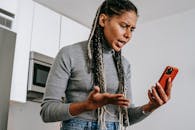 A young woman expressing frustration during a phone call in her kitchen, using a smartphone.
